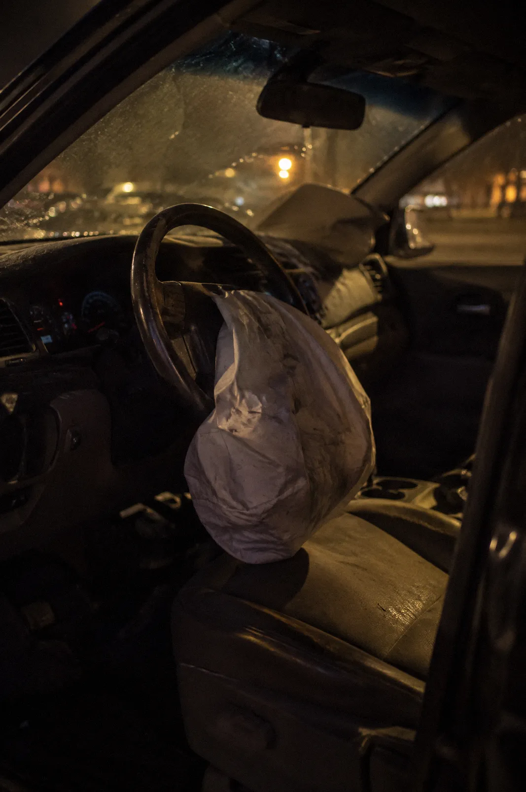 A deflated deployed airbag hanging from a steering wheel, dust in the air, shot through the driver door
