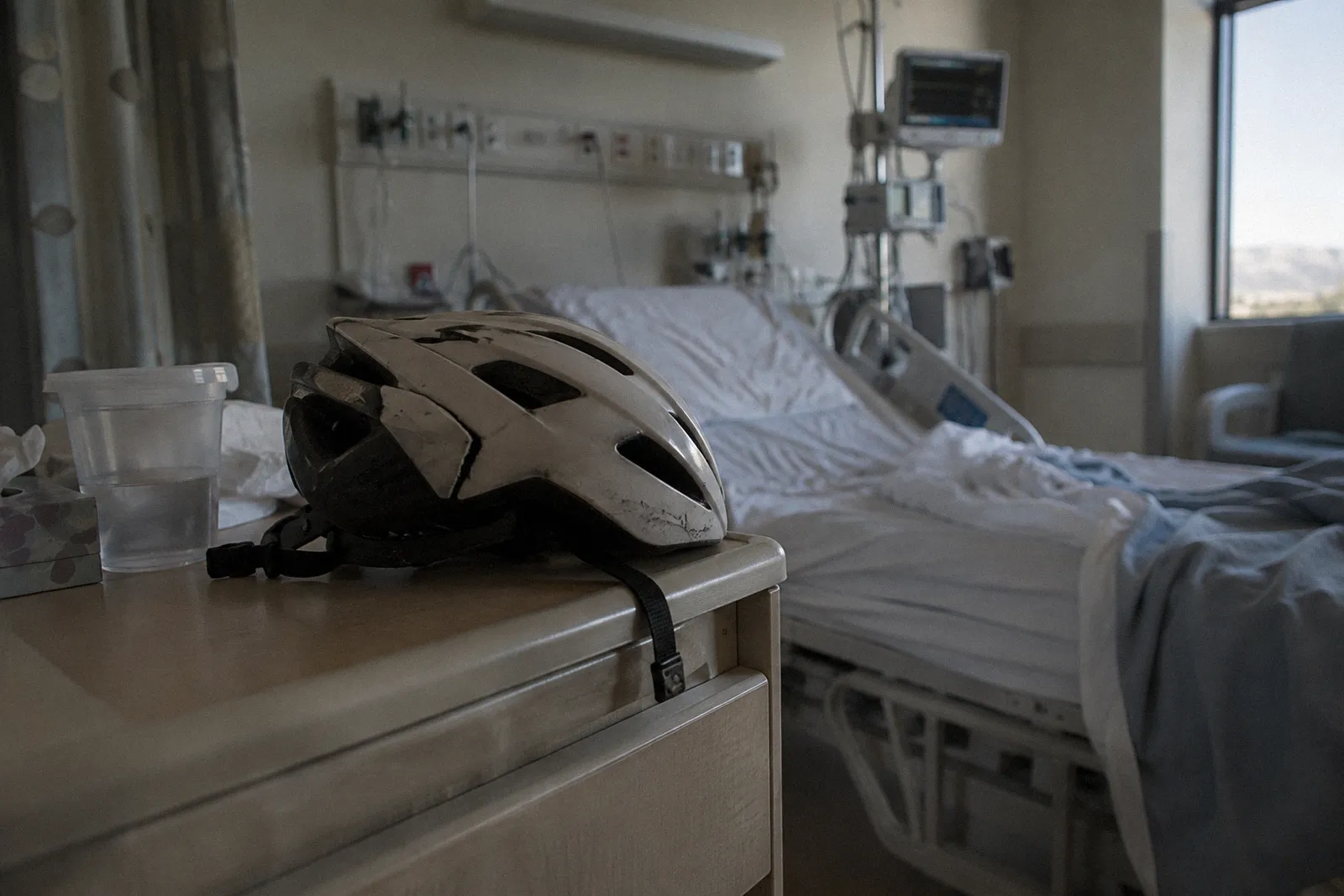 A cracked bicycle helmet resting on a hospital bedside table beside a cup of water