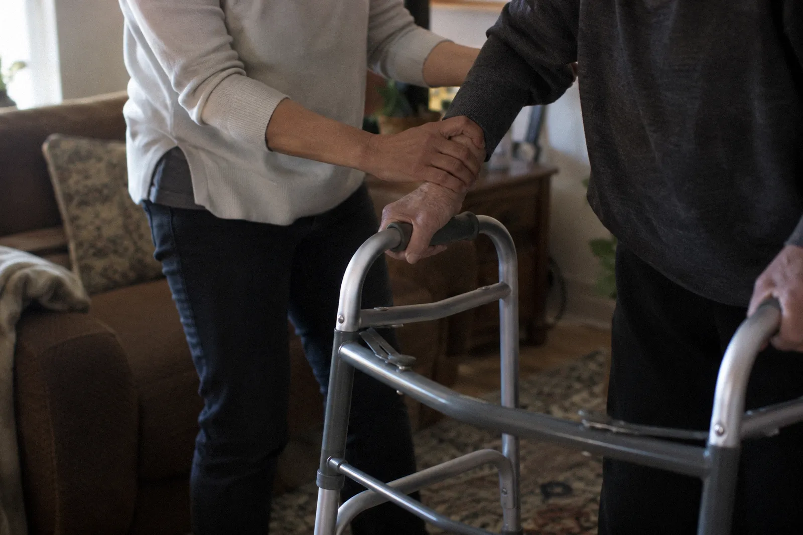 A caregiver's hands steadying another person's forearm on a walker in a living room, faces not shown