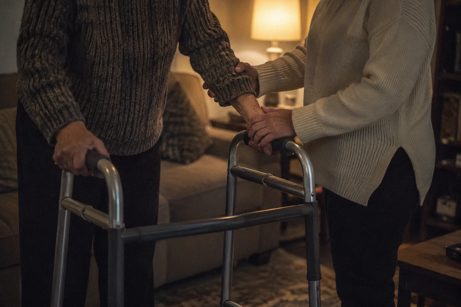A caregiver's hands steadying another person's forearm on a walker in a living room, faces not shown