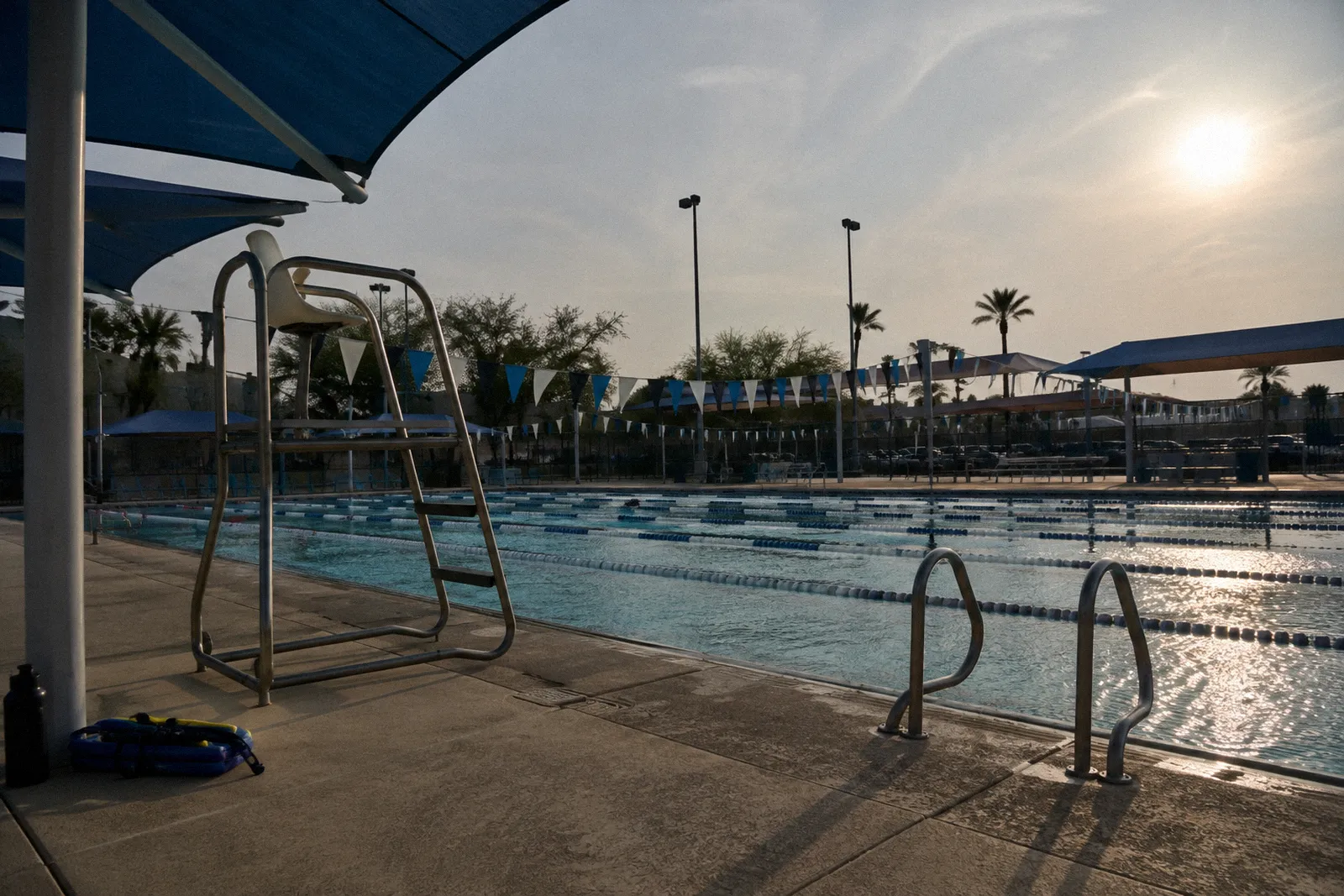 An outdoor public aquatic center in the Phoenix area with lap lanes, an empty lifeguard stand and shade sails, midday