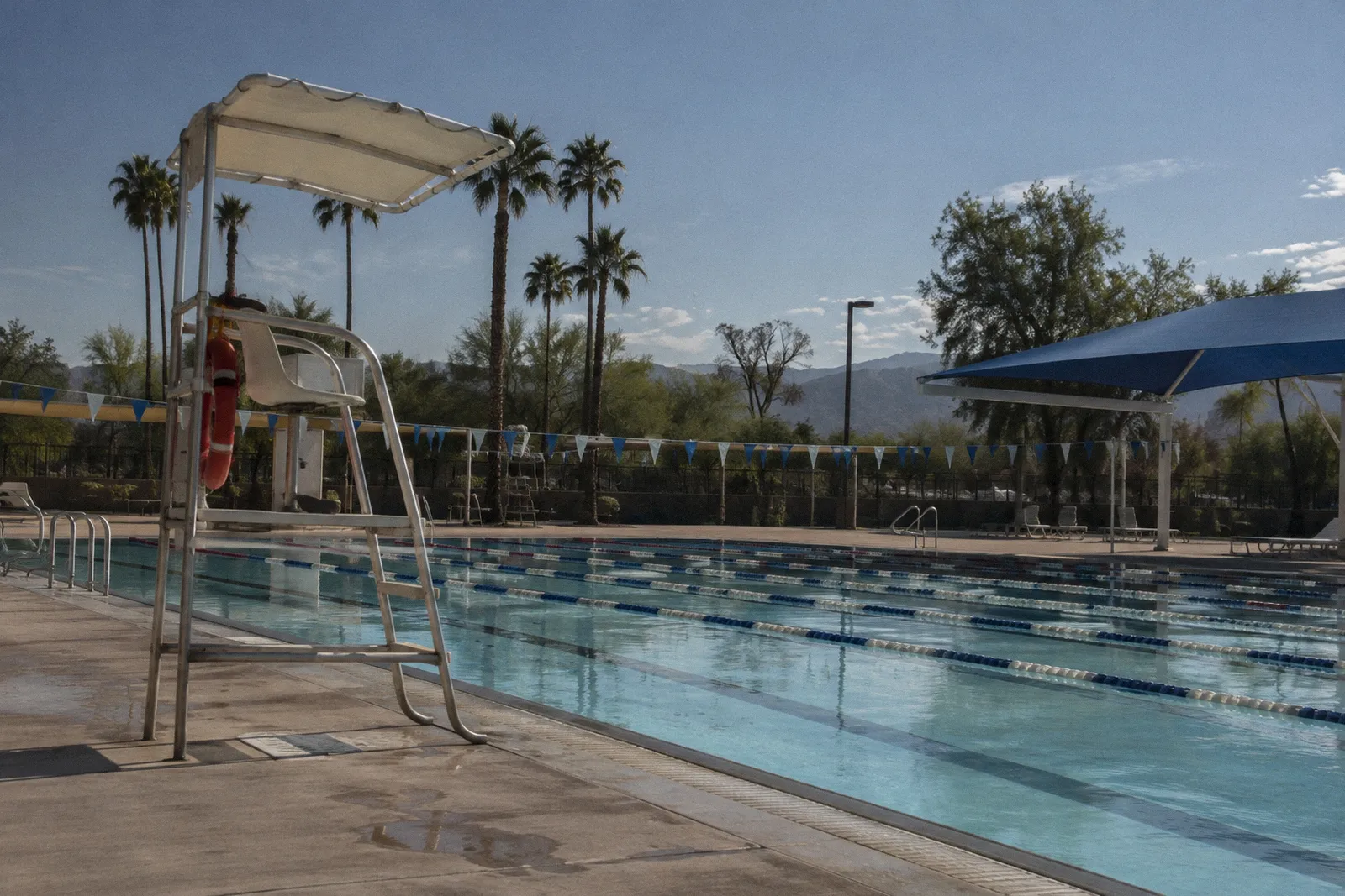 An outdoor public aquatic center in the Phoenix area with lap lanes, an empty lifeguard stand and shade sails, midday