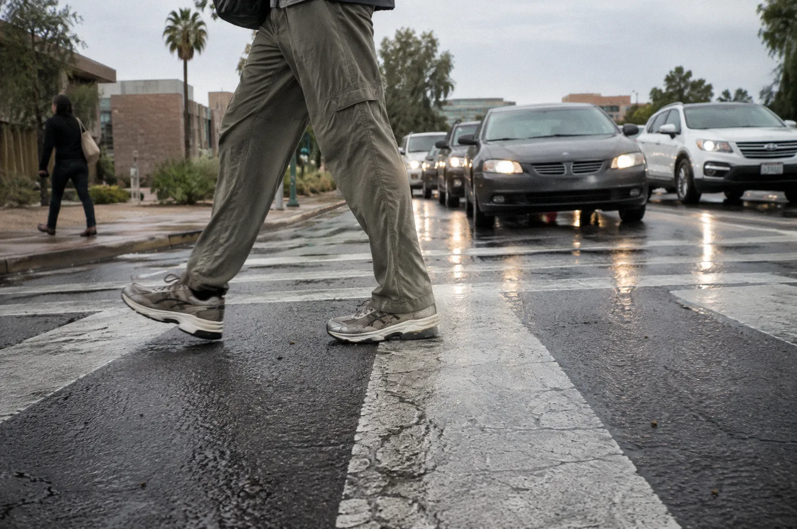 A pedestrian's legs and shoes mid-stride in a painted crosswalk, low angle, cars stopped behind the stop bar