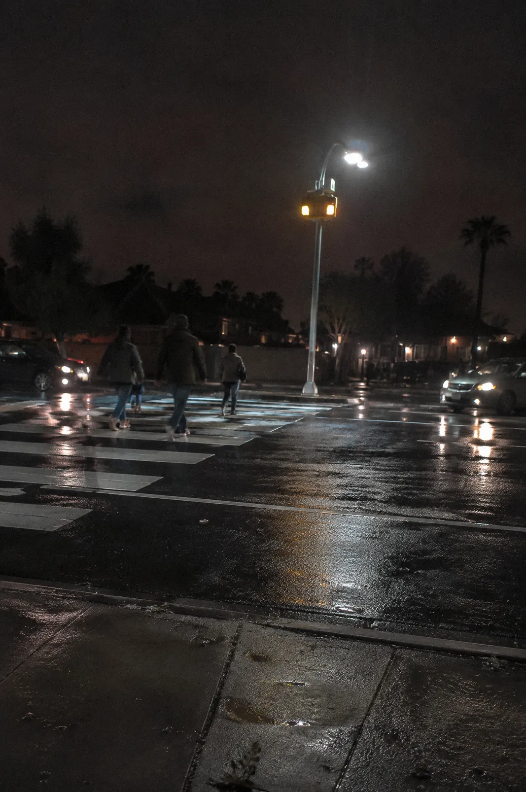 A mid-block crosswalk at night lit by a pedestrian beacon, wet pavement reflections after a storm