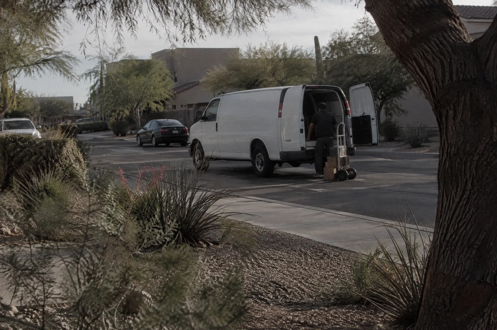An unmarked white delivery van double-parked on a residential street, a hand truck by the rear door