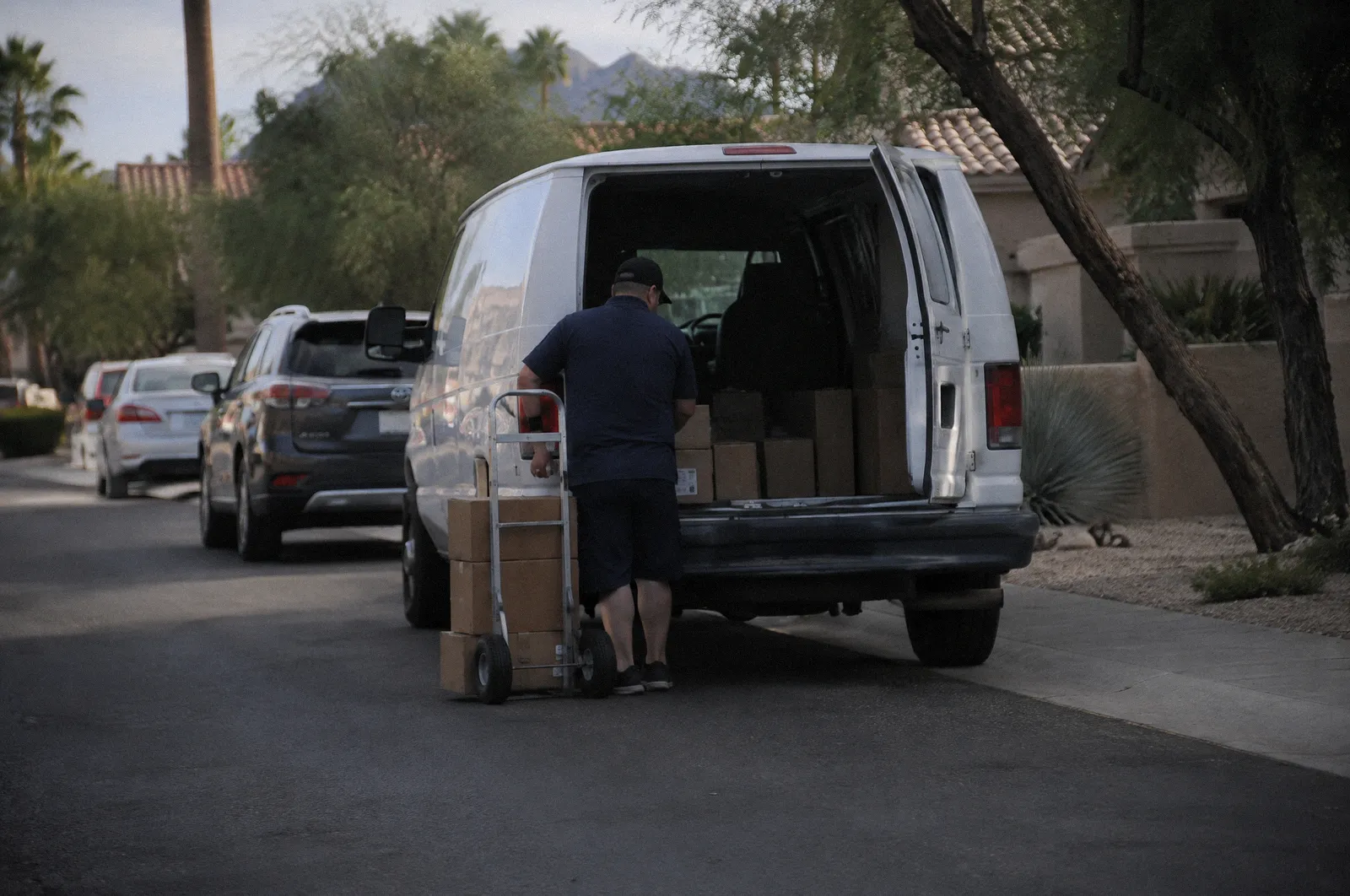 An unmarked white delivery van double-parked on a residential street, a hand truck by the rear door