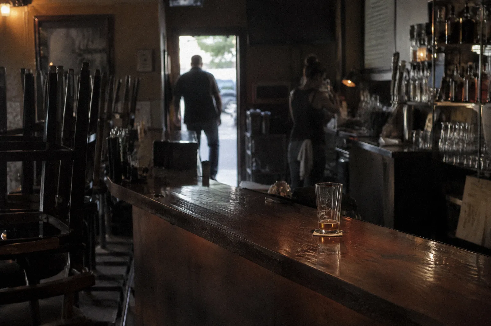The interior of a dim bar at closing time, stools up, a single glass on the counter, no readable signs