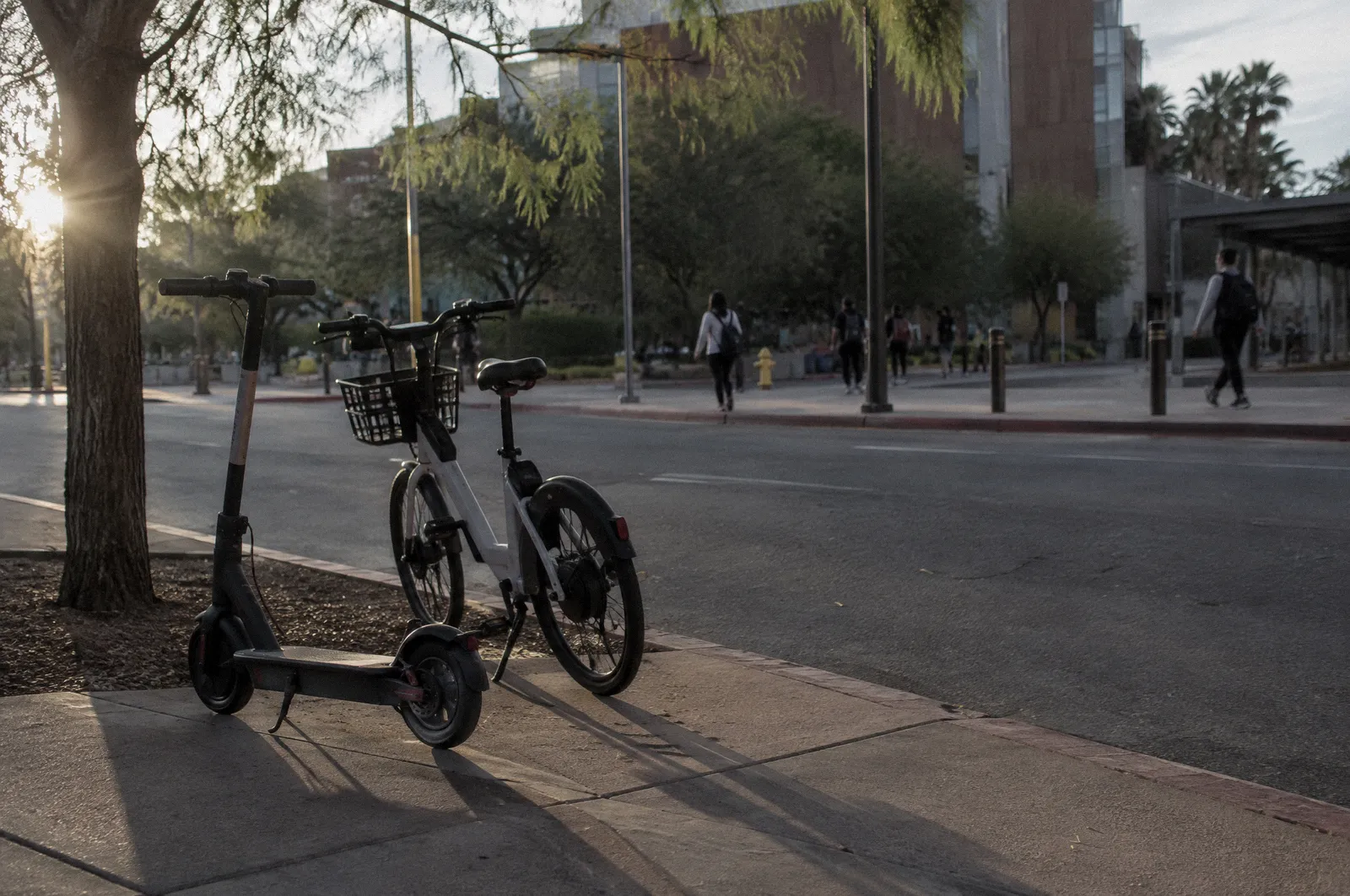 A rental e-scooter and an e-bike leaning by a downtown Tempe curb, students far in the background
