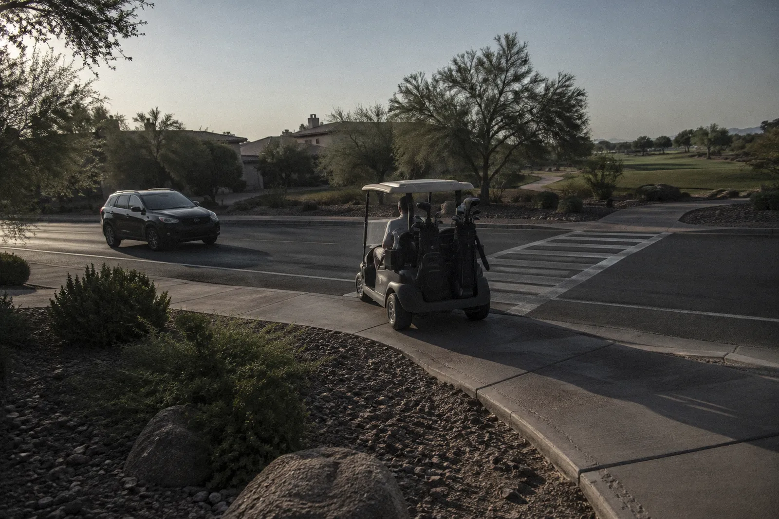 A golf cart path crossing a two-lane neighborhood road, painted crossing lines, a cart waiting to cross