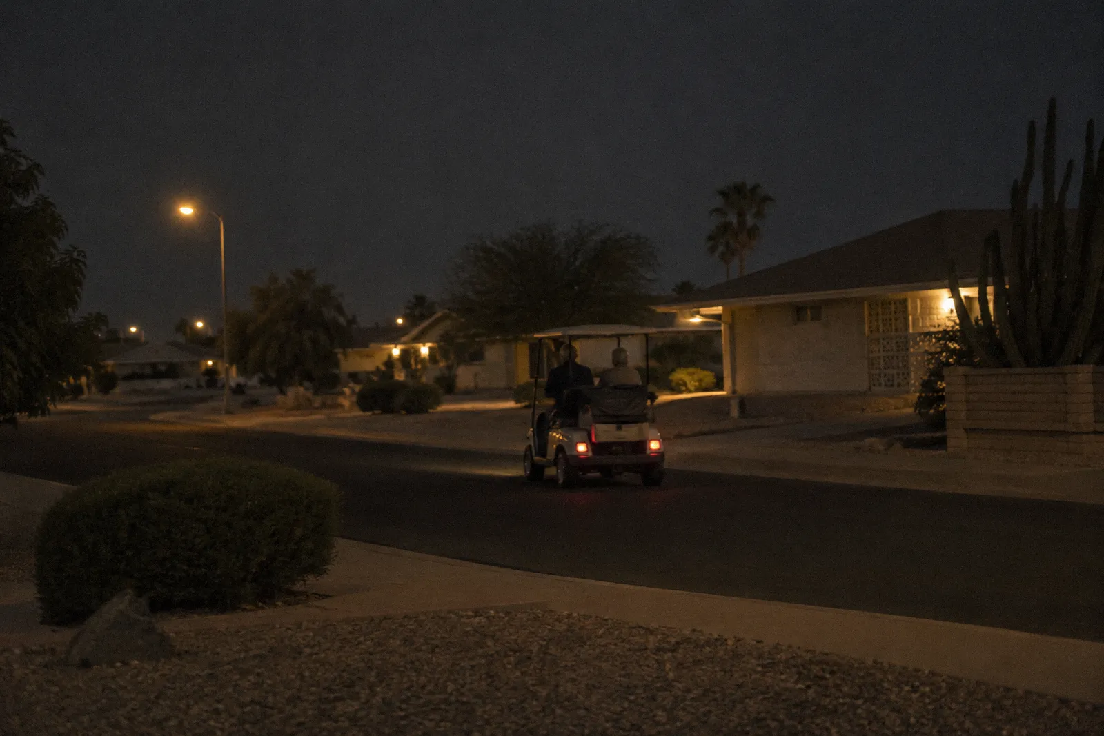 A single golf cart driving along a quiet residential street in a Sun City style retirement community, low homes and gravel yards