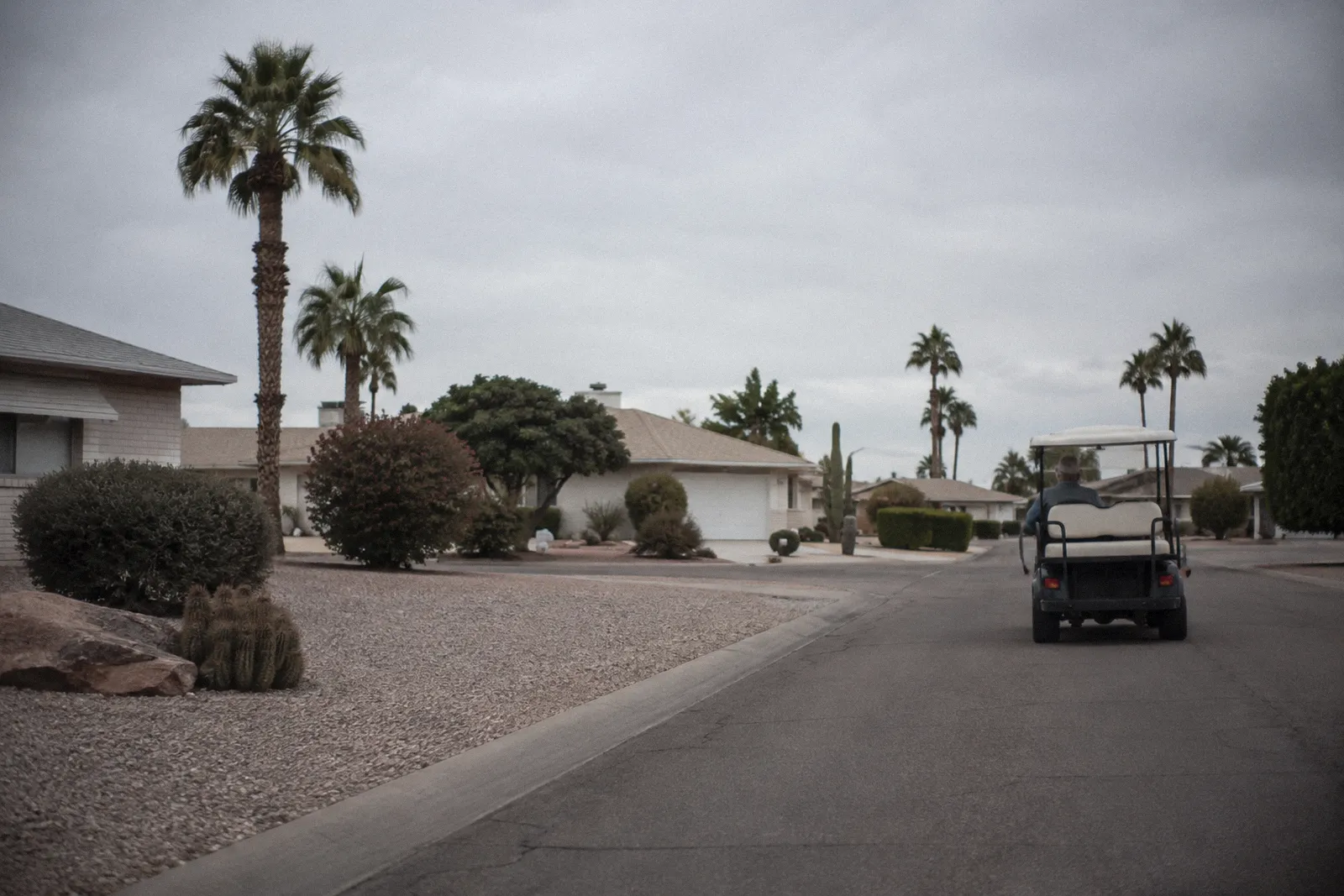 A single golf cart driving along a quiet residential street in a Sun City style retirement community, low homes and gravel yards