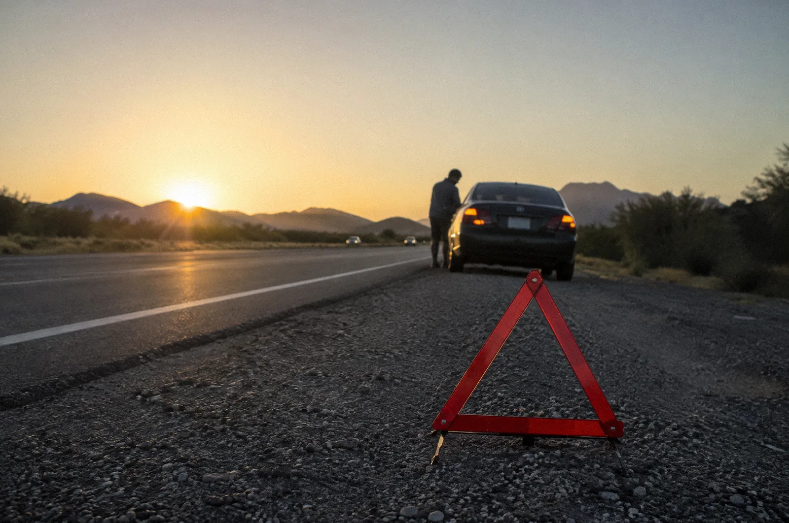 A red reflective warning triangle on a road shoulder behind a stopped car with hazard lights, golden hour