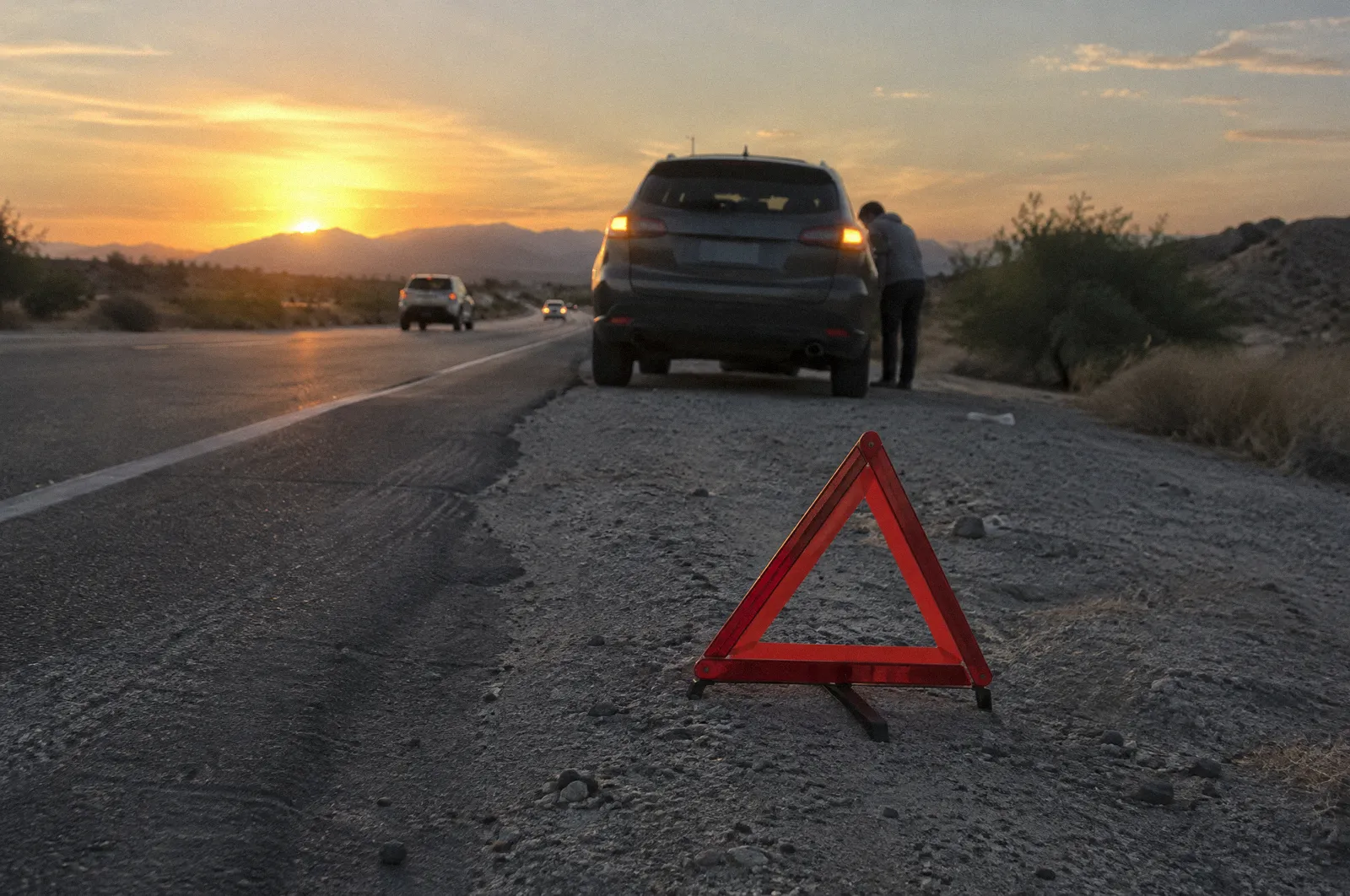 A red reflective warning triangle on a road shoulder behind a stopped car with hazard lights, golden hour