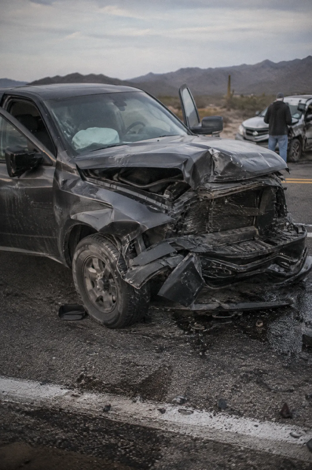 The crushed front end of a pickup truck after a head-on crash on a two-lane desert road, deployed airbag visible