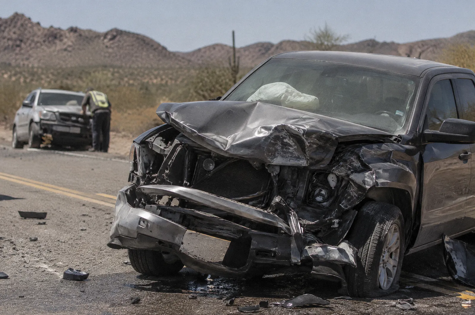 The crushed front end of a pickup truck after a head-on crash on a two-lane desert road, deployed airbag visible