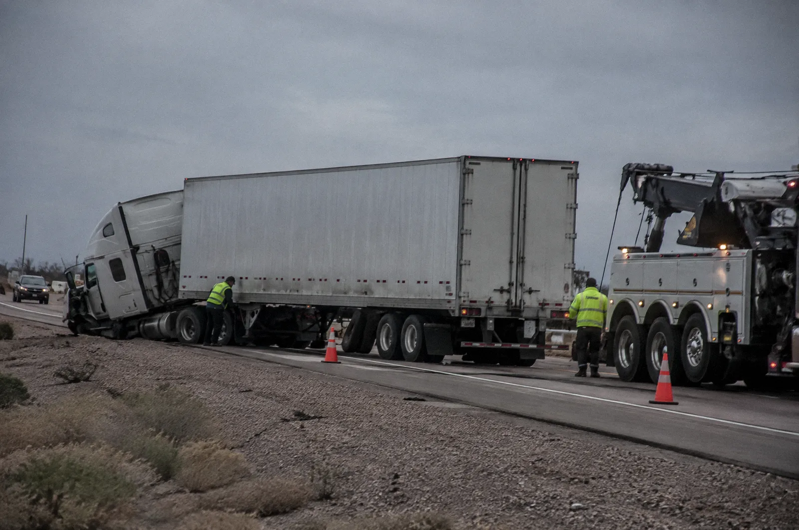 A jackknifed semi trailer on a freeway shoulder after a crash, cones and a tow rig, overcast