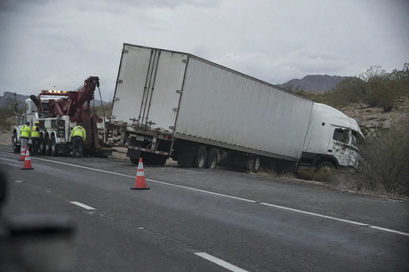 A jackknifed semi trailer on a freeway shoulder after a crash, cones and a tow rig, overcast