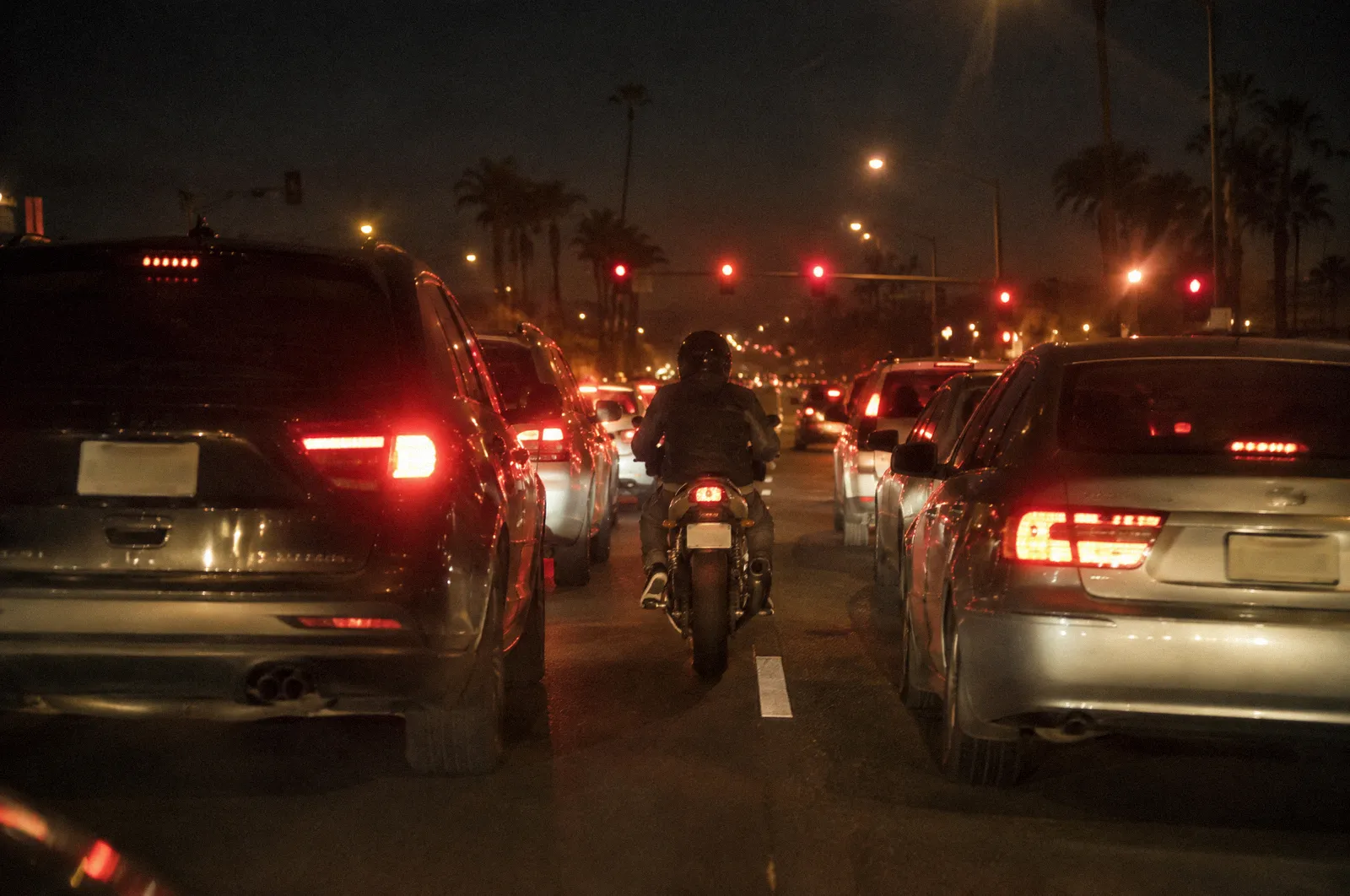 A motorcycle moving slowly between two lanes of fully stopped cars at a Phoenix intersection, shot from behind
