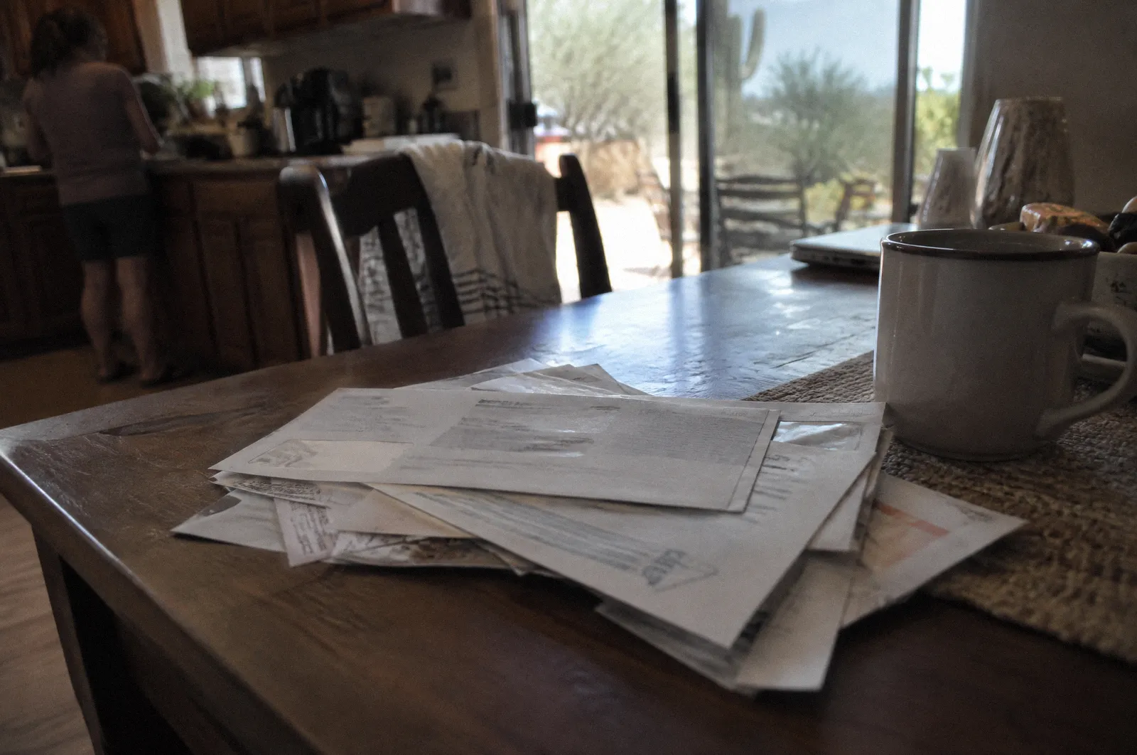 A stack of hospital bills and envelopes on a kitchen table, all text blurred and unreadable, coffee mug