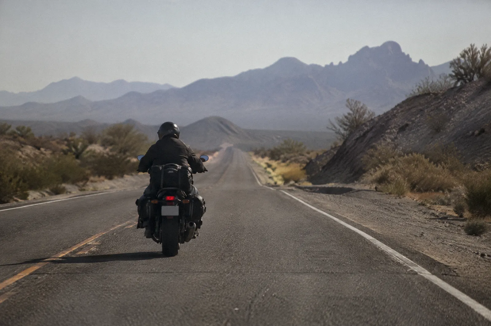A motorcycle rider from behind on an empty desert highway, helmet and jacket, heat shimmer