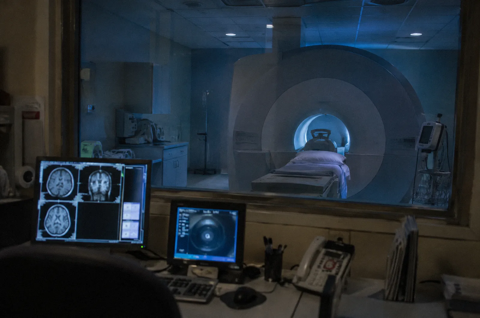 An MRI scanner room seen through a control room window, soft blue light, nobody in frame