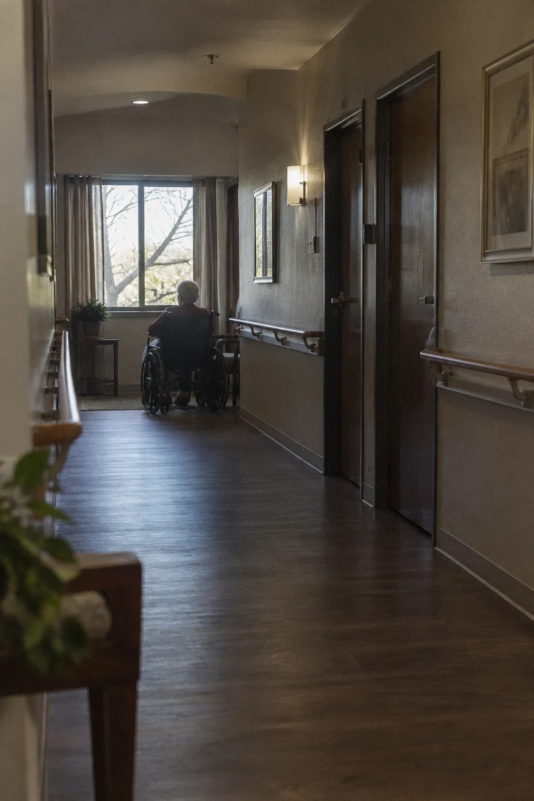 A quiet assisted living hallway with wooden handrails, closed doors and a wheelchair parked by a window
