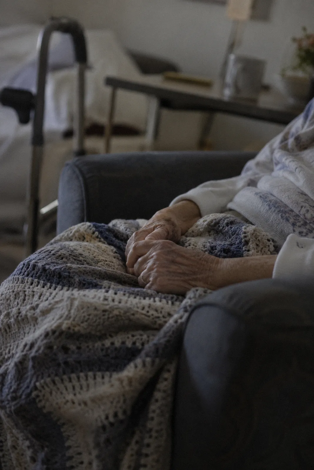 An elderly person's hands resting on a knit blanket in a care facility chair, face not shown