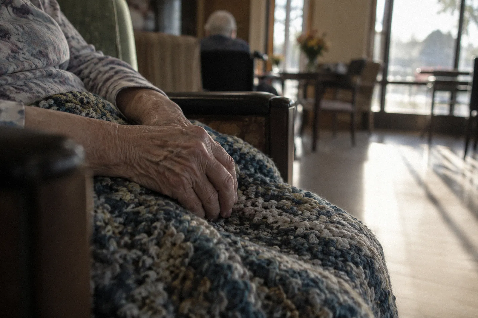An elderly person's hands resting on a knit blanket in a care facility chair, face not shown