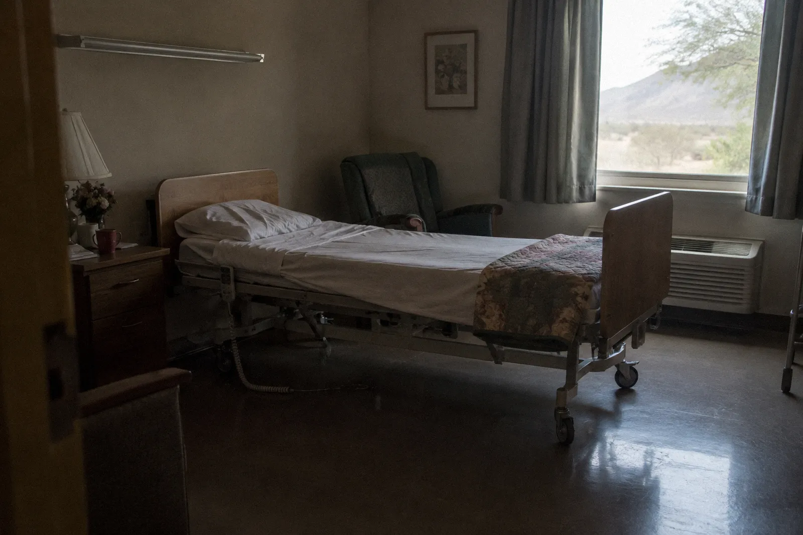 An empty care facility room with an adjustable bed, a call button cord, a folded blanket and window light