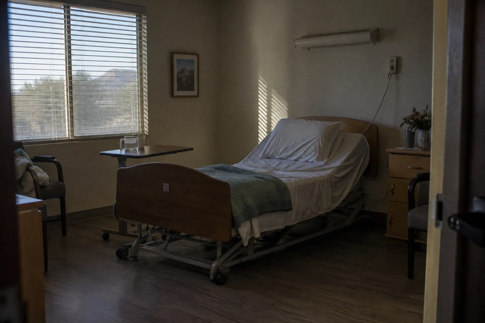 An empty care facility room with an adjustable bed, a call button cord, a folded blanket and window light
