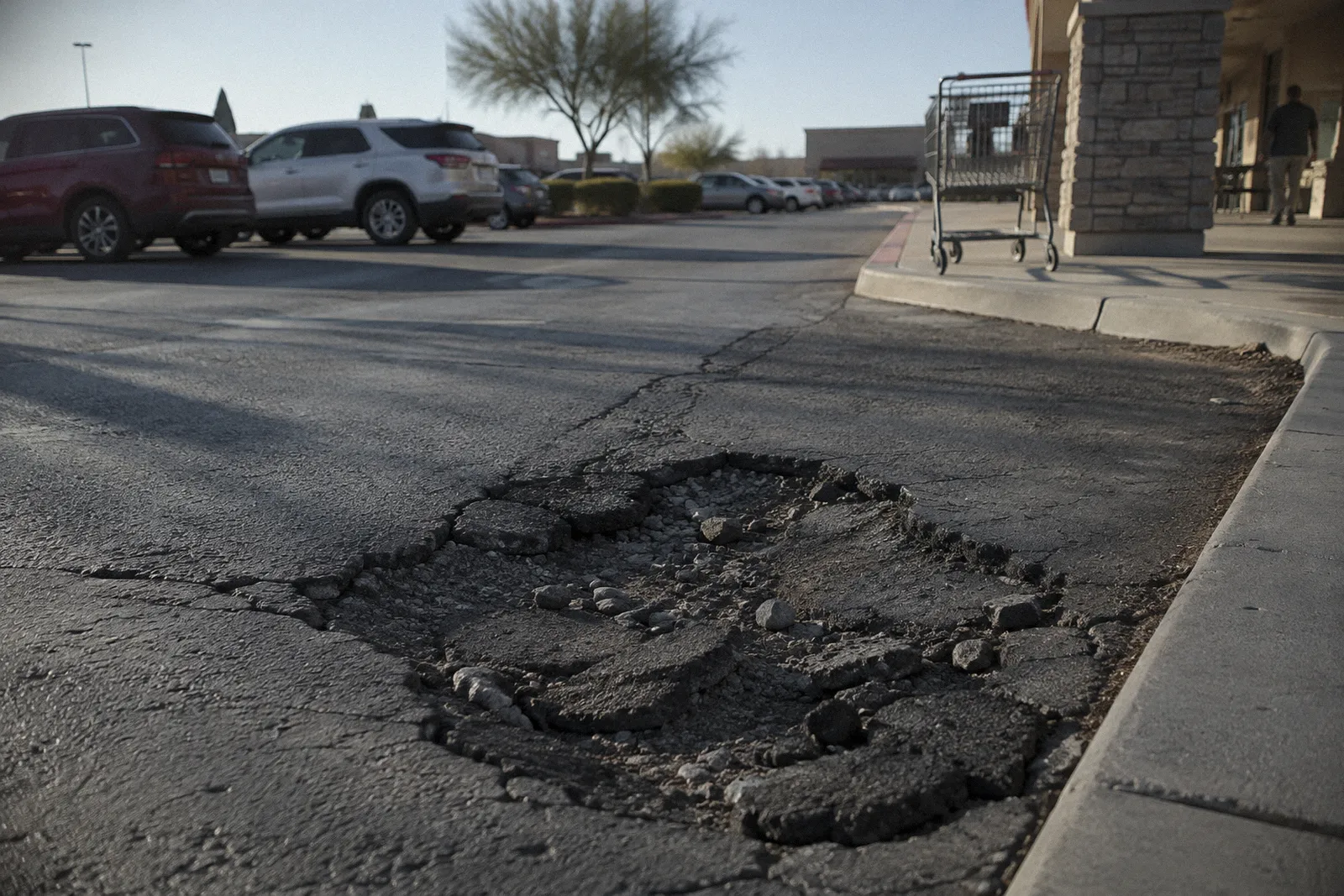 A deep pothole near a parking lot walkway at a strip mall, a shopping cart in the background, afternoon