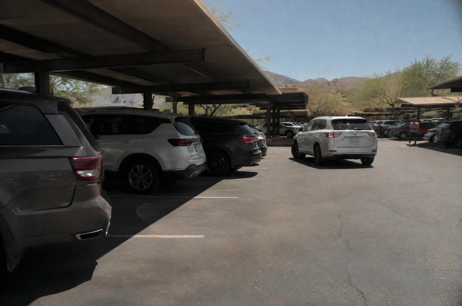A sun-baked shopping center parking lot with rows of cars, shade structures, a car reversing out