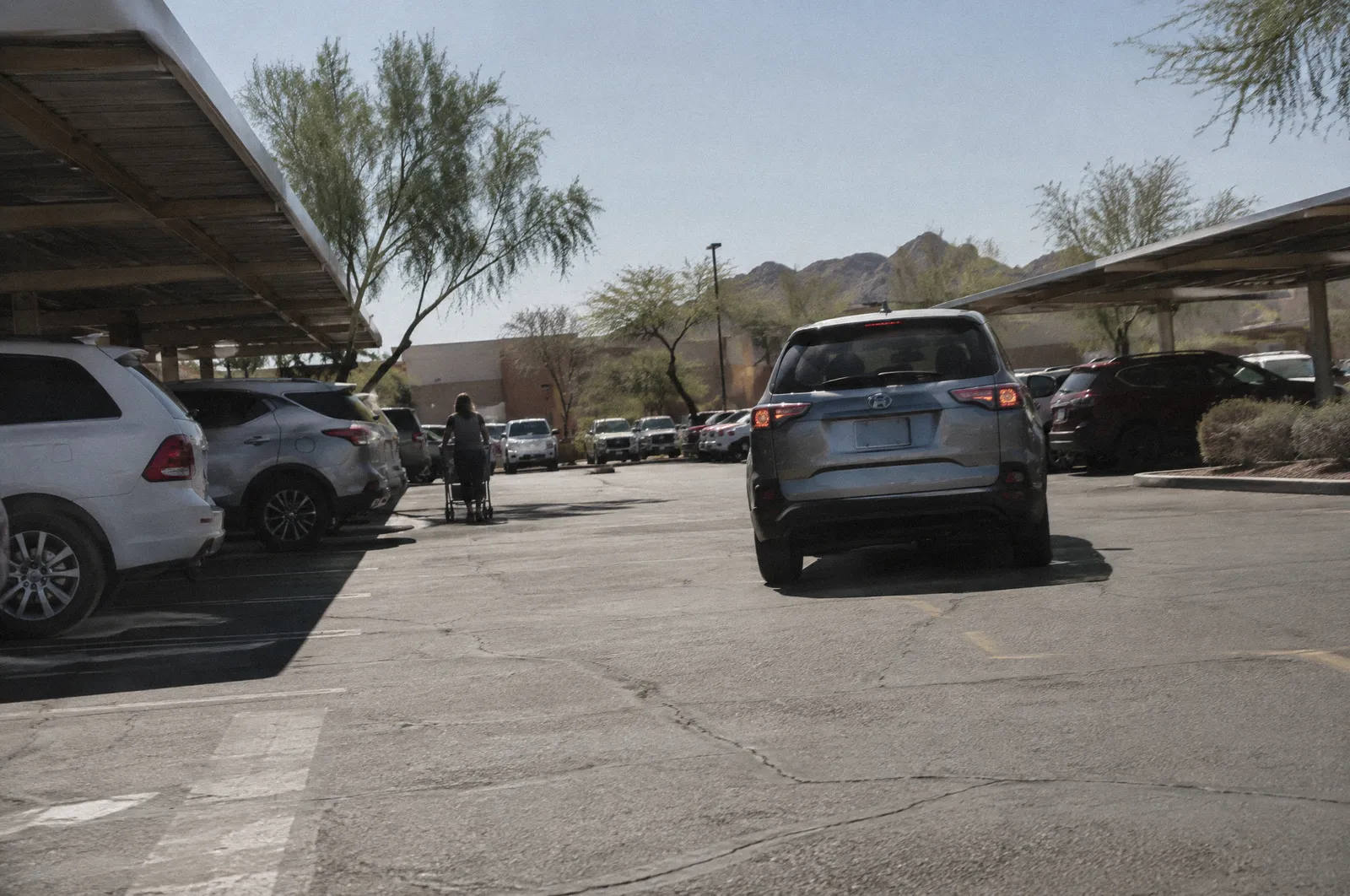 A sun-baked shopping center parking lot with rows of cars, shade structures, a car reversing out