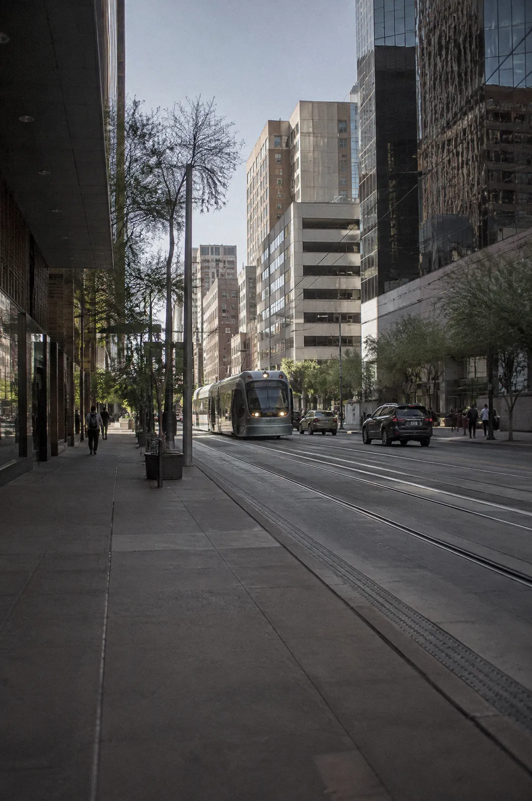 A downtown Phoenix street canyon at midday, light rail tracks in the pavement, pedestrians far away as small figures