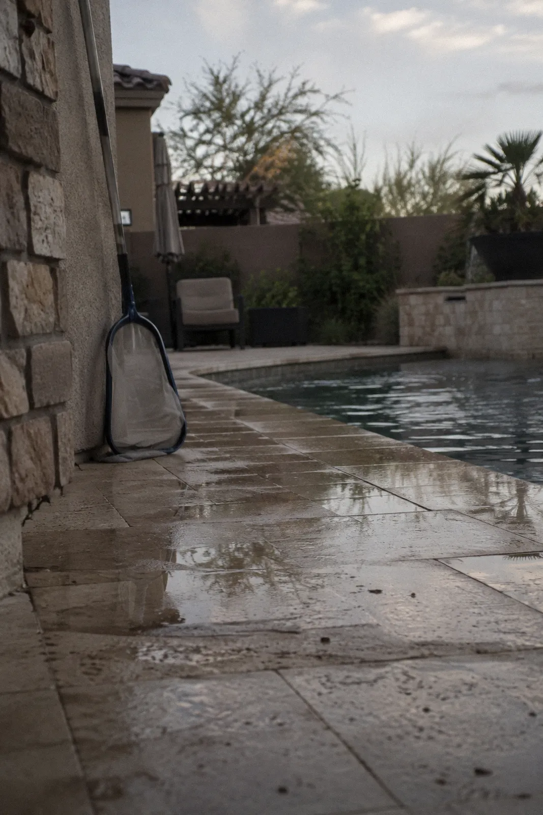 Wet travertine tiles at the edge of a backyard pool with a puddle and a pool skimmer net leaning on the wall