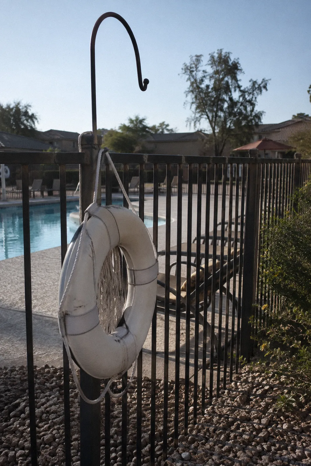 A white life ring and a shepherd's hook mounted on a fence beside a community pool, harsh shadows