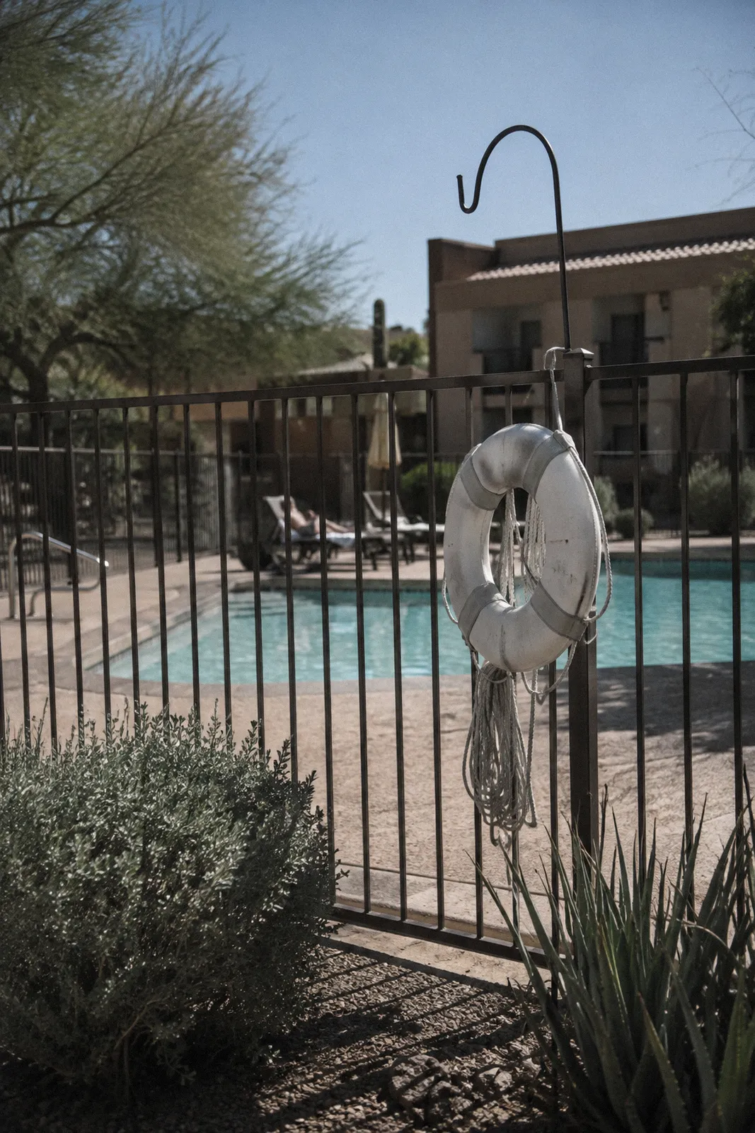 A white life ring and a shepherd's hook mounted on a fence beside a community pool, harsh shadows