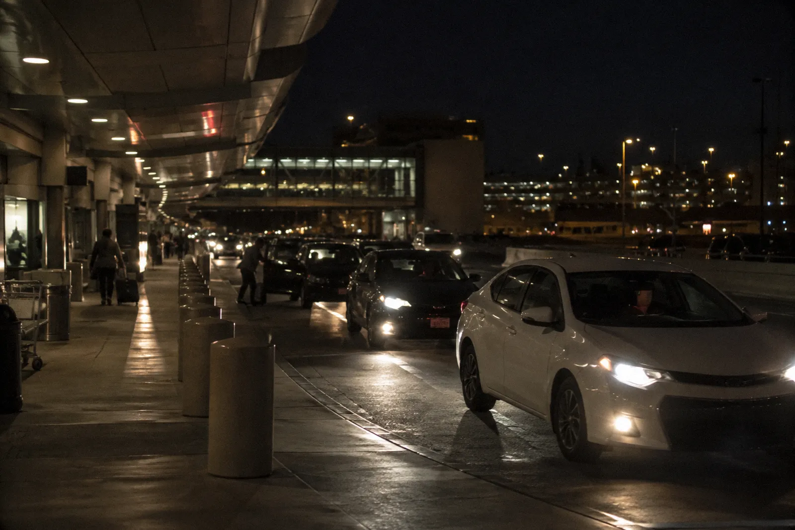 A rideshare pickup curb at a Phoenix airport terminal at night, cars waiting, travelers far away, no readable signs