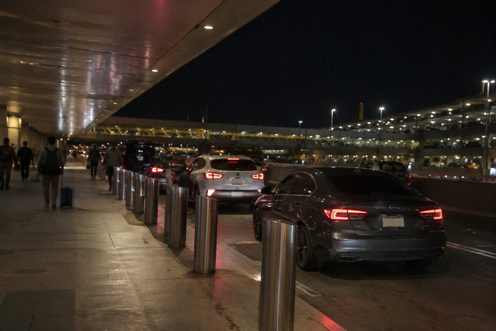 A rideshare pickup curb at a Phoenix airport terminal at night, cars waiting, travelers far away, no readable signs