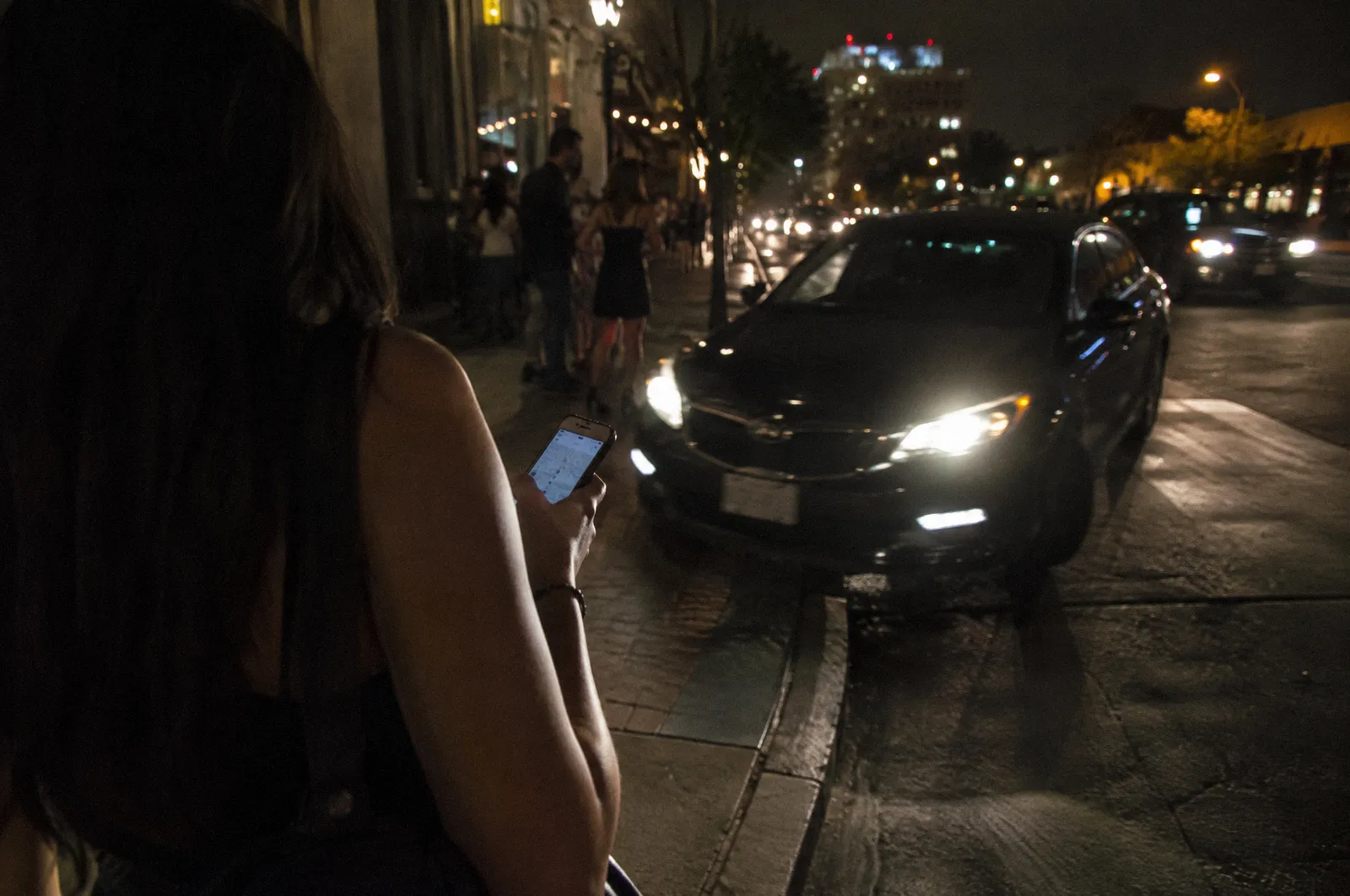 A car pulling to the curb for a pickup outside a nightlife district at night, a phone in a hand in the foreground