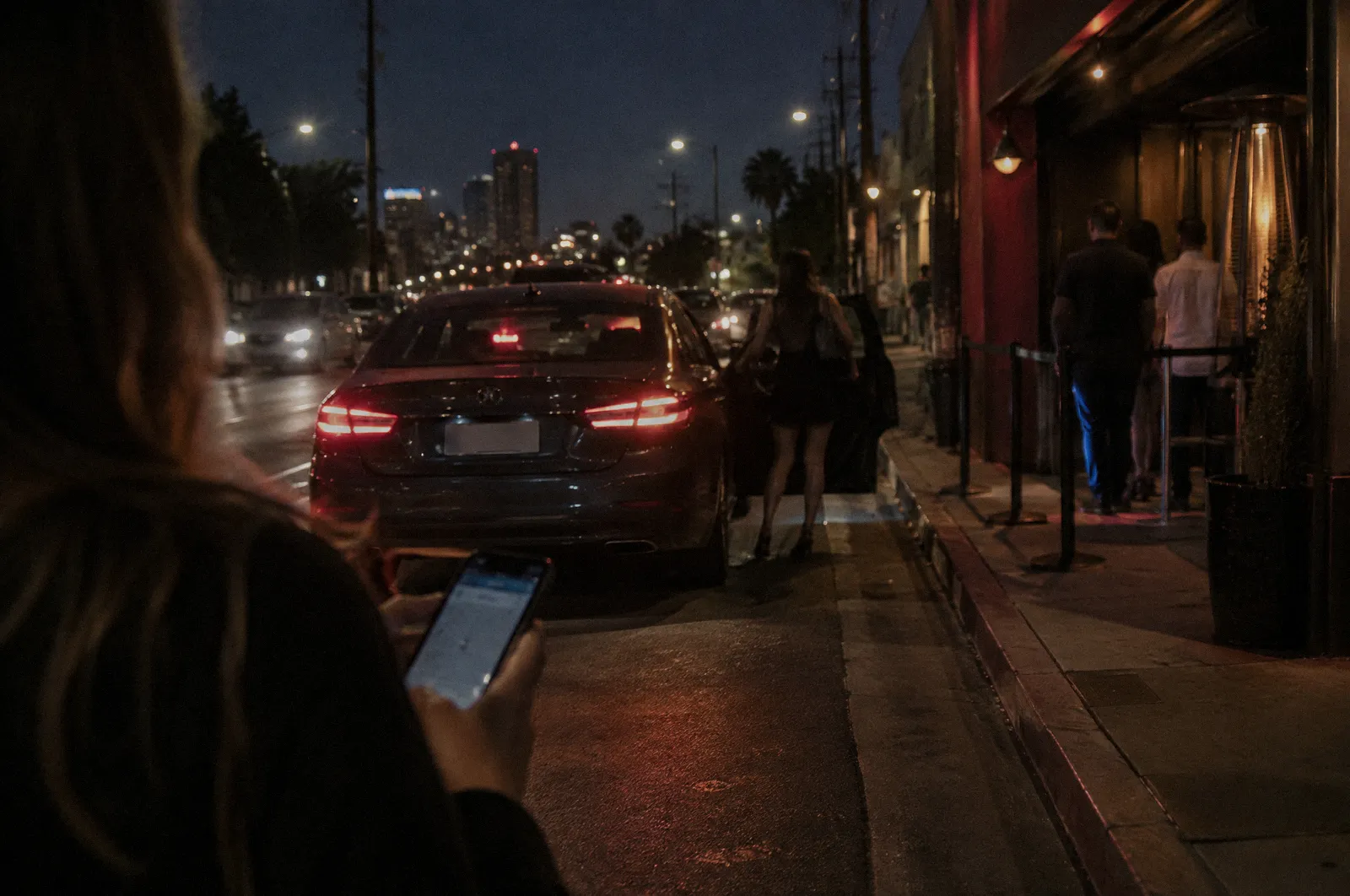 A car pulling to the curb for a pickup outside a nightlife district at night, a phone in a hand in the foreground