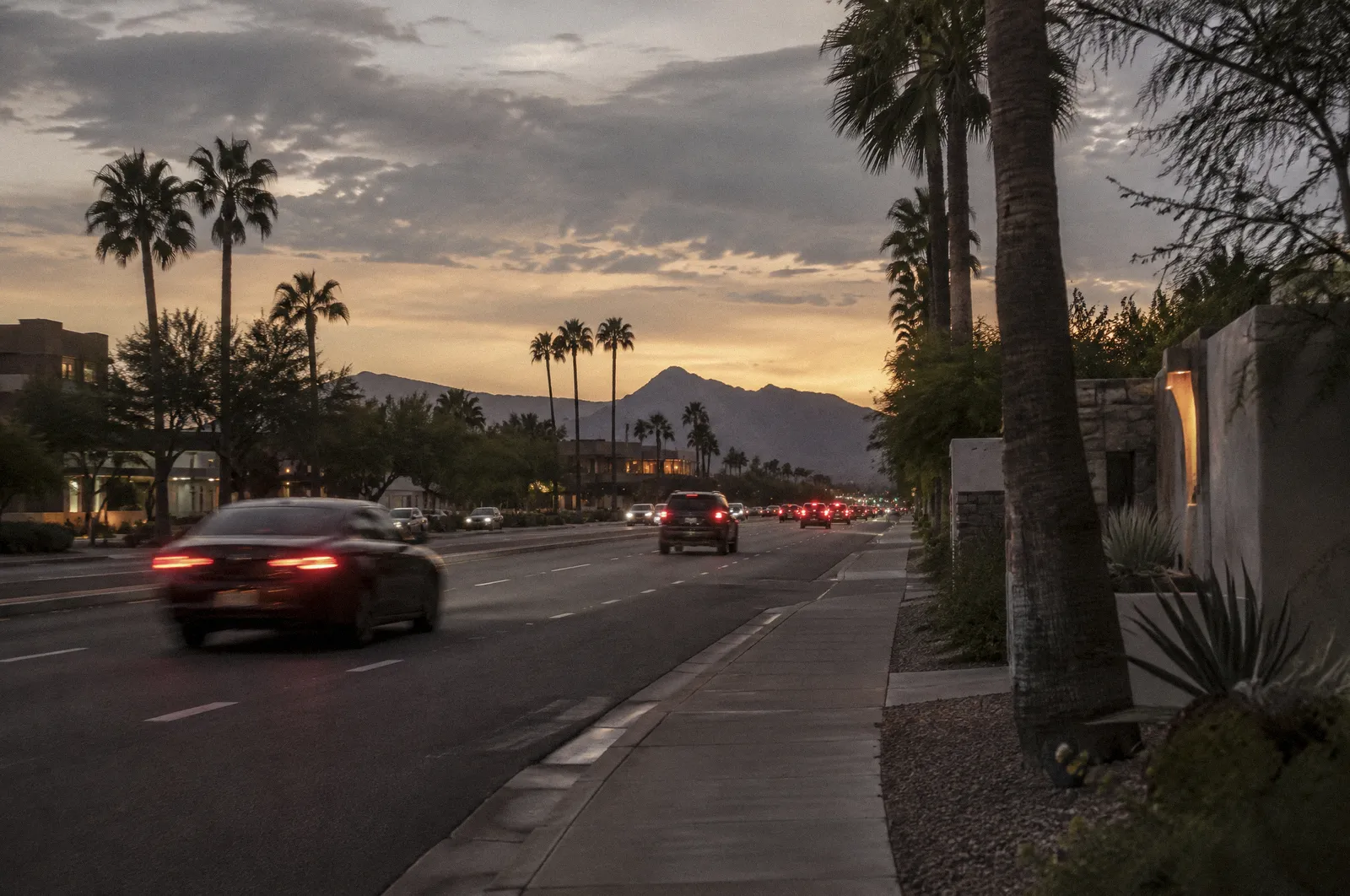 Scottsdale Road at dusk seen from a sidewalk, palm trees, light traffic with motion-blurred taillights, McDowell Mountains faint on the horizon
