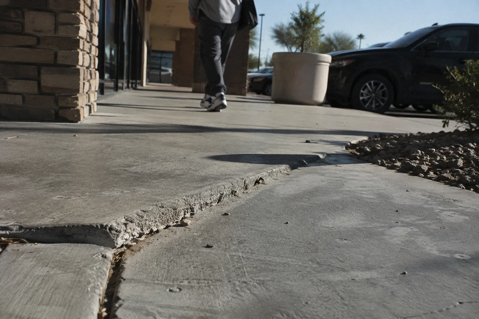 A lifted, cracked section of concrete sidewalk forming a trip edge in a shopping center walkway, harsh shadow