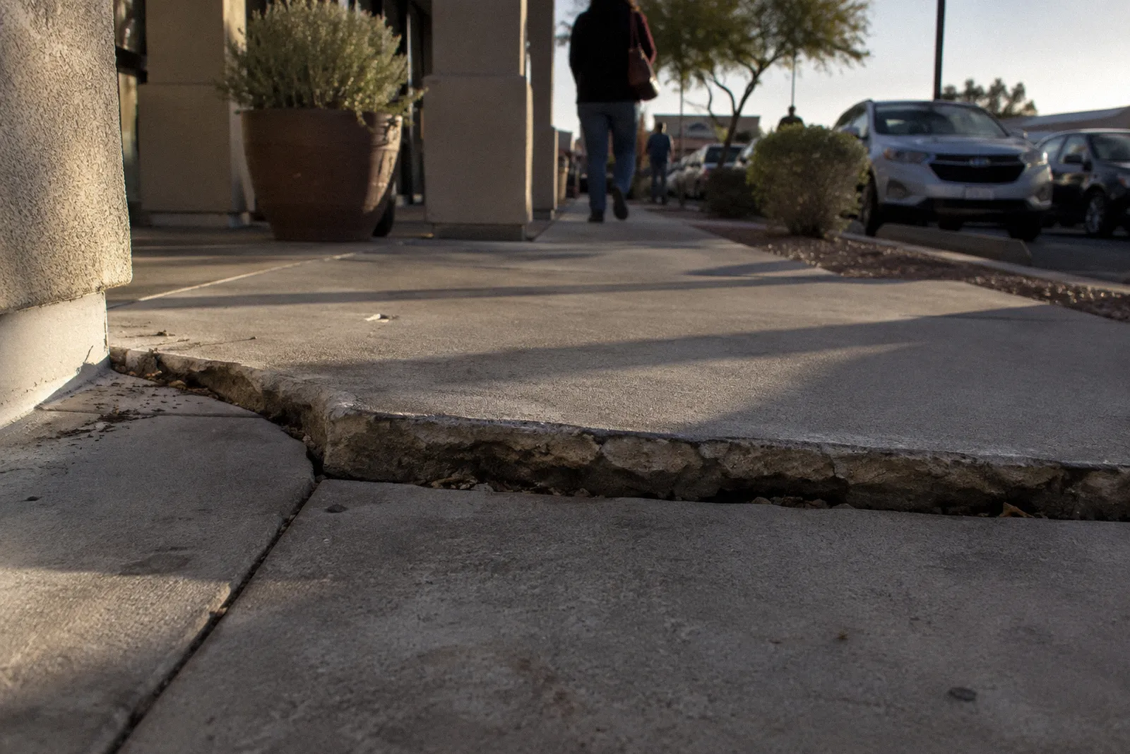 A lifted, cracked section of concrete sidewalk forming a trip edge in a shopping center walkway, harsh shadow