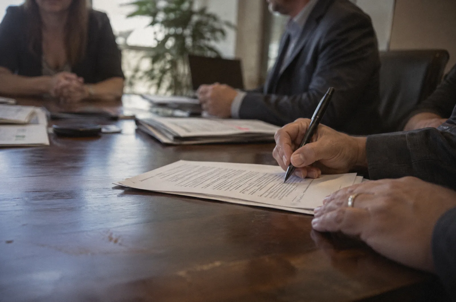 Hands signing a document at a conference table, the text unreadable, warm office light