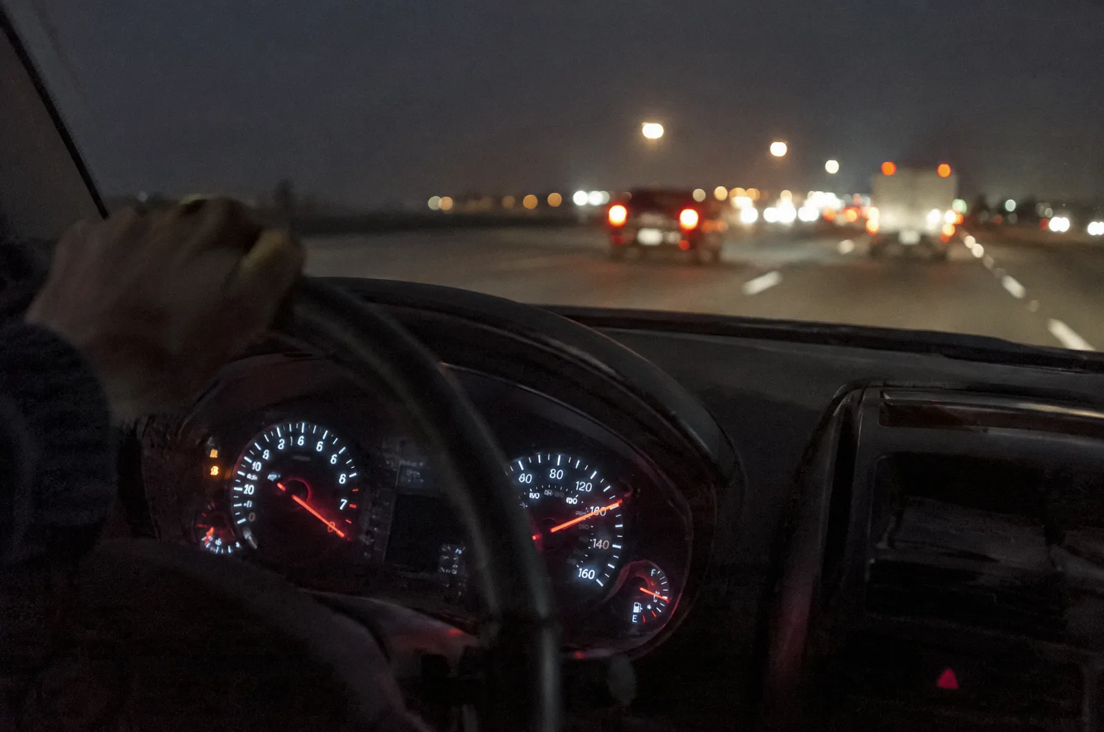 A car instrument cluster at speed at night, needle high, the road ahead blurred through the windshield