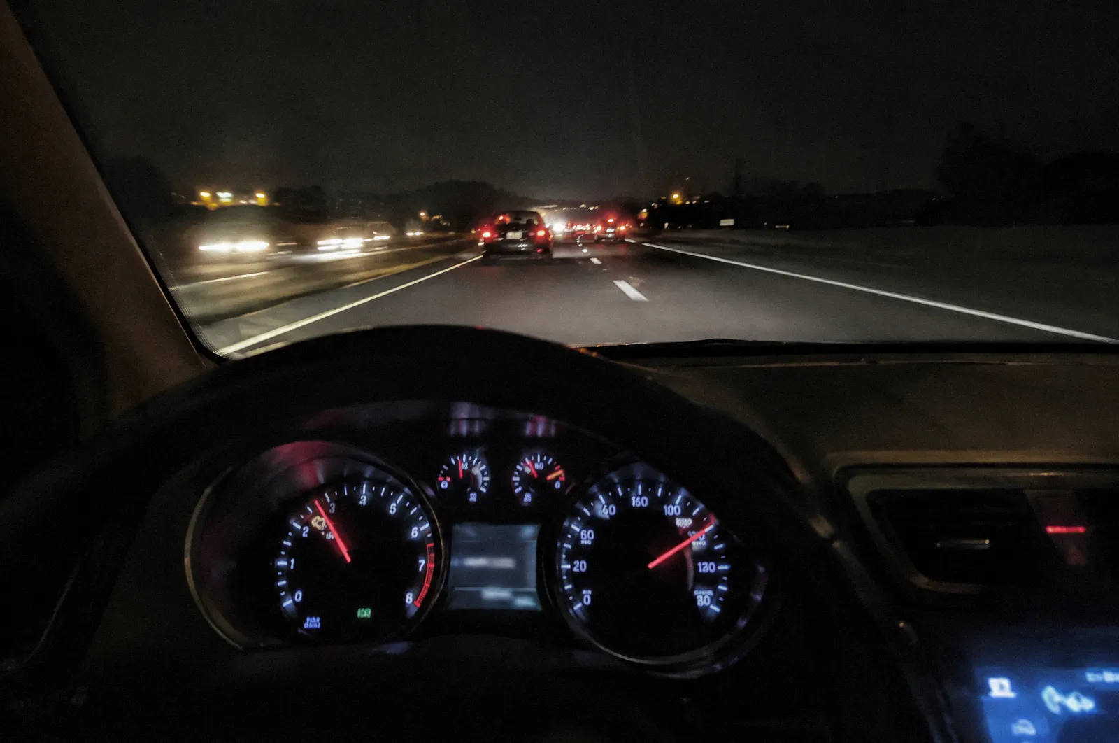 A car instrument cluster at speed at night, needle high, the road ahead blurred through the windshield