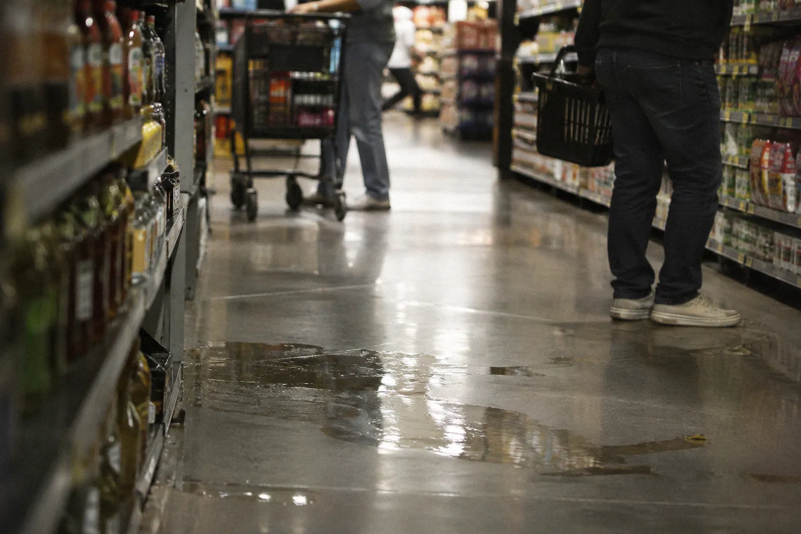 A grocery store aisle with a clear liquid spill on the polished floor, shelves blurred, no readable labels