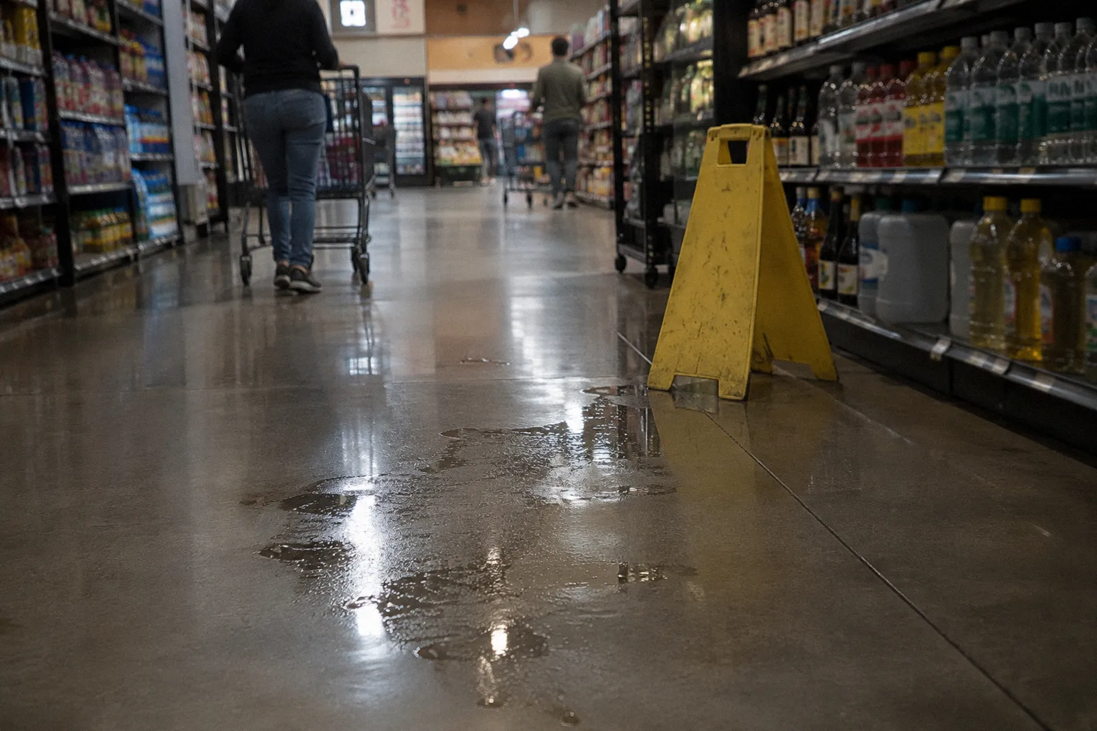 A grocery store aisle with a clear liquid spill on the polished floor, shelves blurred, no readable labels