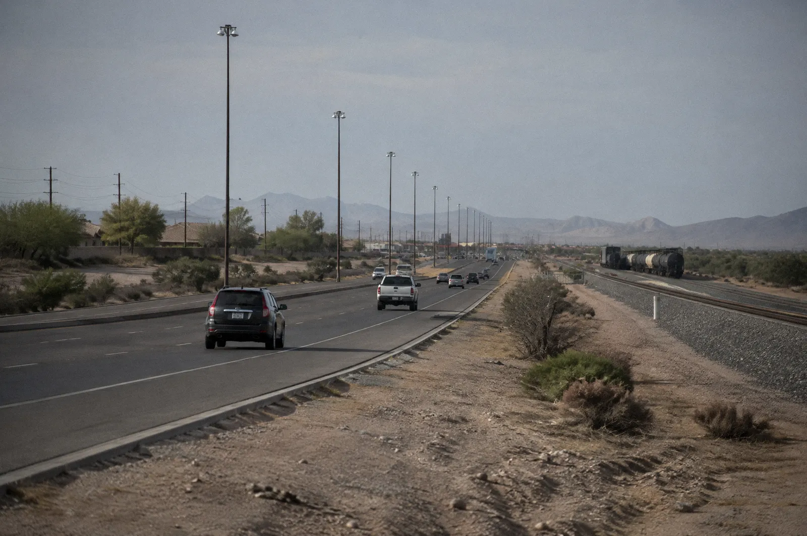 A long diagonal highway through the far West Valley near Surprise with rail tracks alongside, flat light